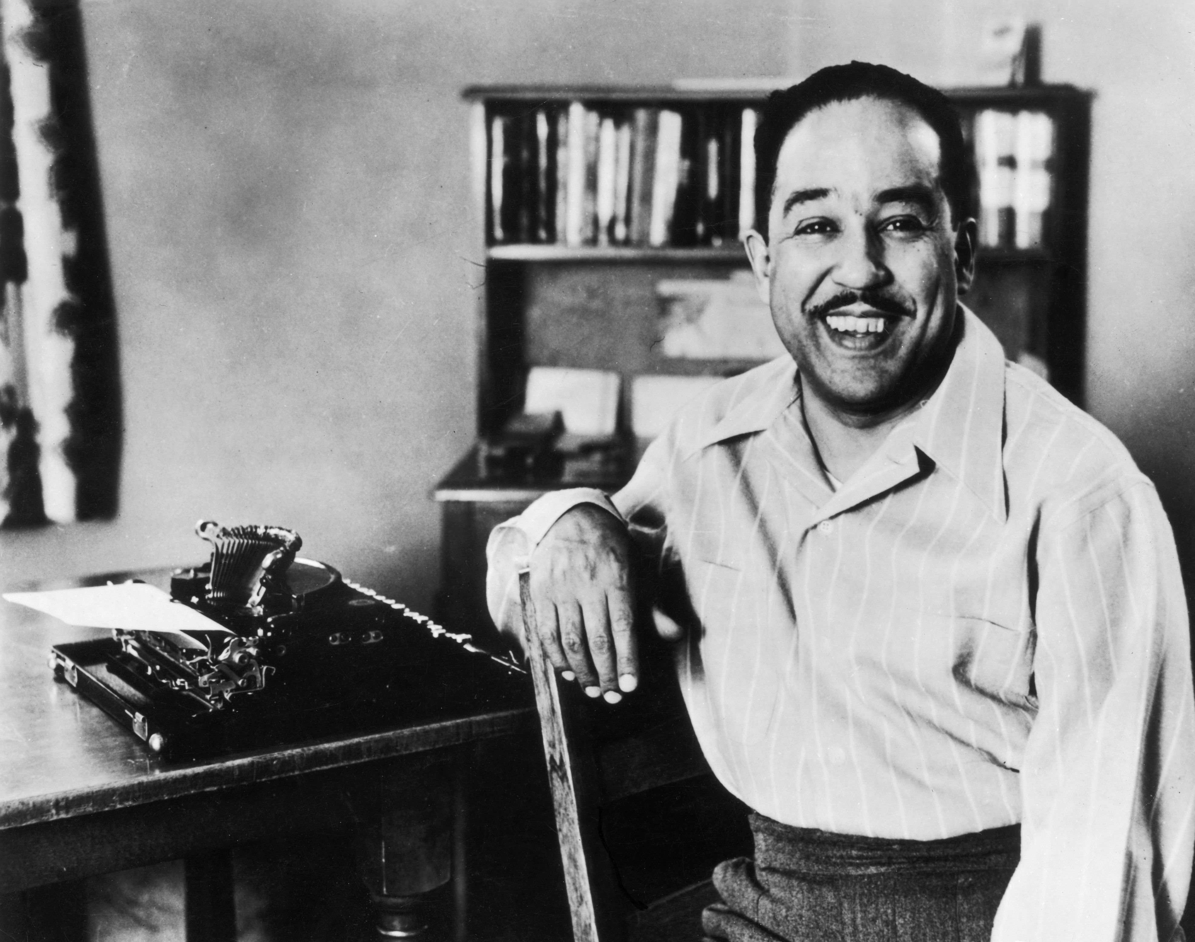 Langston Hughes sonríe alegre en una silla al lado de una máquina de escribir con una hoja de papel sobre una mesa. Al fondo se ve una biblioteca.