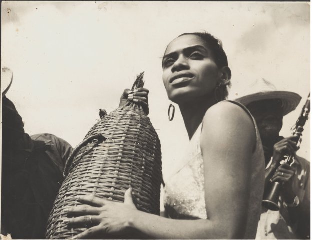 Fotografía en blanco y negro de Delia Zapata Olivella con aretes y pelo recogido ayuda a sostener una artesanía de fibra vegetal a un hombre con sombrero. Detrás de ella un hombre afro con sombrero sostiene un clarinete.