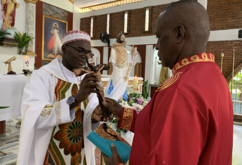 El p. Elías Libanda imc, delegado de Pastoral Afro Cali, entrega el crucifijo a Ferneyz Carabalí, mientras este laico misionero de la Consolata sostiene una biblia. Al fondo se ve la imagen de Cristo Resucitado en el templo de Cristo Maestro.